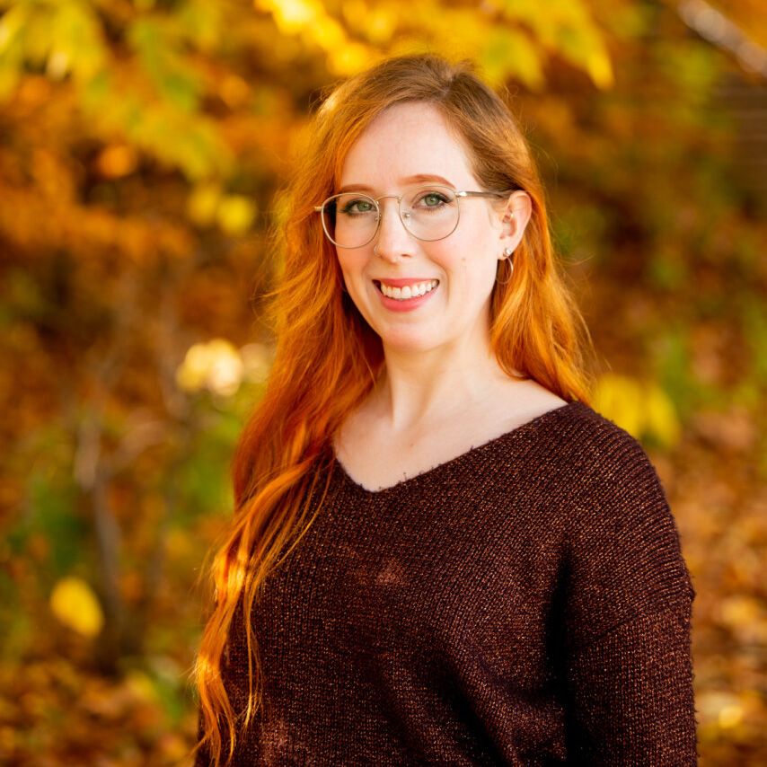 a young woman with long red hair facing the camera with autumn foliage in the background