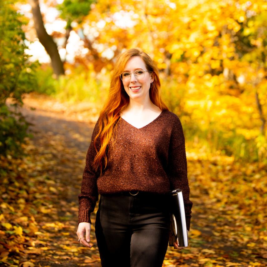 a young woman with long red hair walking towards the camera with autumn foliage in the background